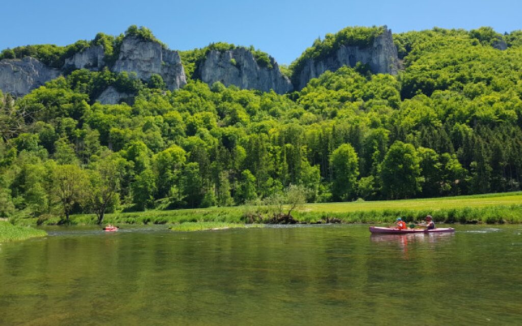 Themenbild von Kanufahrern auf der Donau im Hintergrund sind saftig grüne Wiesen, grüne Bäume und Felsen vor strahlend blauem Himmel zu sehen