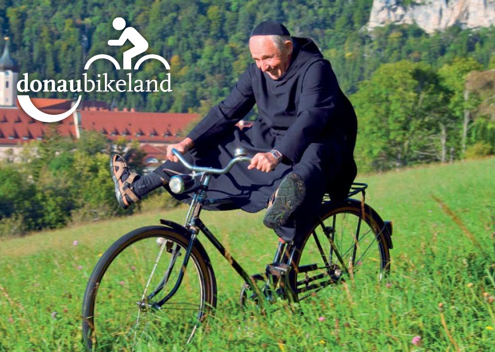 Bild von Mönch auf, der auf einer Wiese Fahrrad fährt und die Beine von sich streckt
