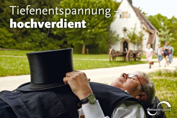 Donaubergland Postkartenmotiv mit schlafendem Mann mit Zylinder auf dem Bauch vor einer Kapelle.