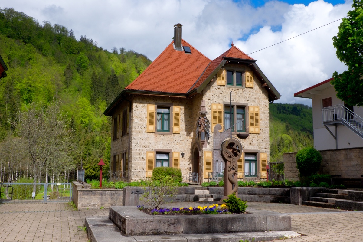 Bild vom Michaelsplatz mit bepflanztem Steinbeet und dem Pfarrhaus mit gelben Fensterläden vor einem bewaldeten Hang in Bärenthal