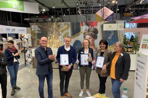 Foto von einer Zertifikatübergabe auf der Messe mit drei Personen mit Zertifikat in der Hand und weiteren Personen, die applaudieren