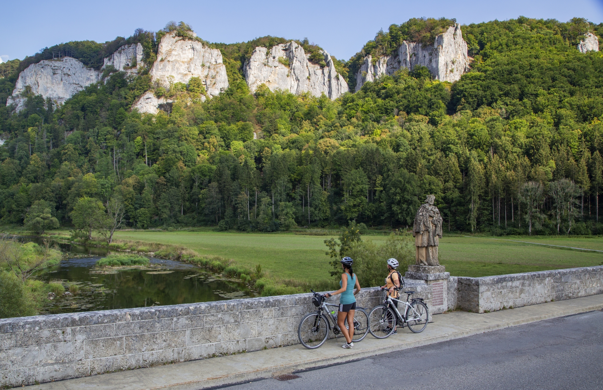 Bild von zwei Radfahrerinnen, die einer Steinmauer stehen und auf die Donau blicken im Hintergrund sind Felsen, Bäume und Wiese zu sehen