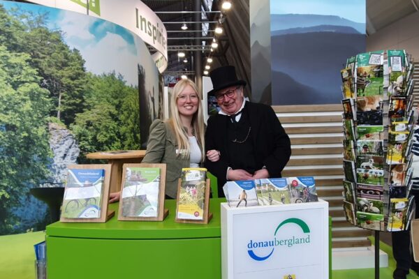 Foto von Stand Donaubergland auf Messe mit Prospekten, Ständer mit Karten und Mann mit Frack und Zylinder und Standmitarbeiterin.