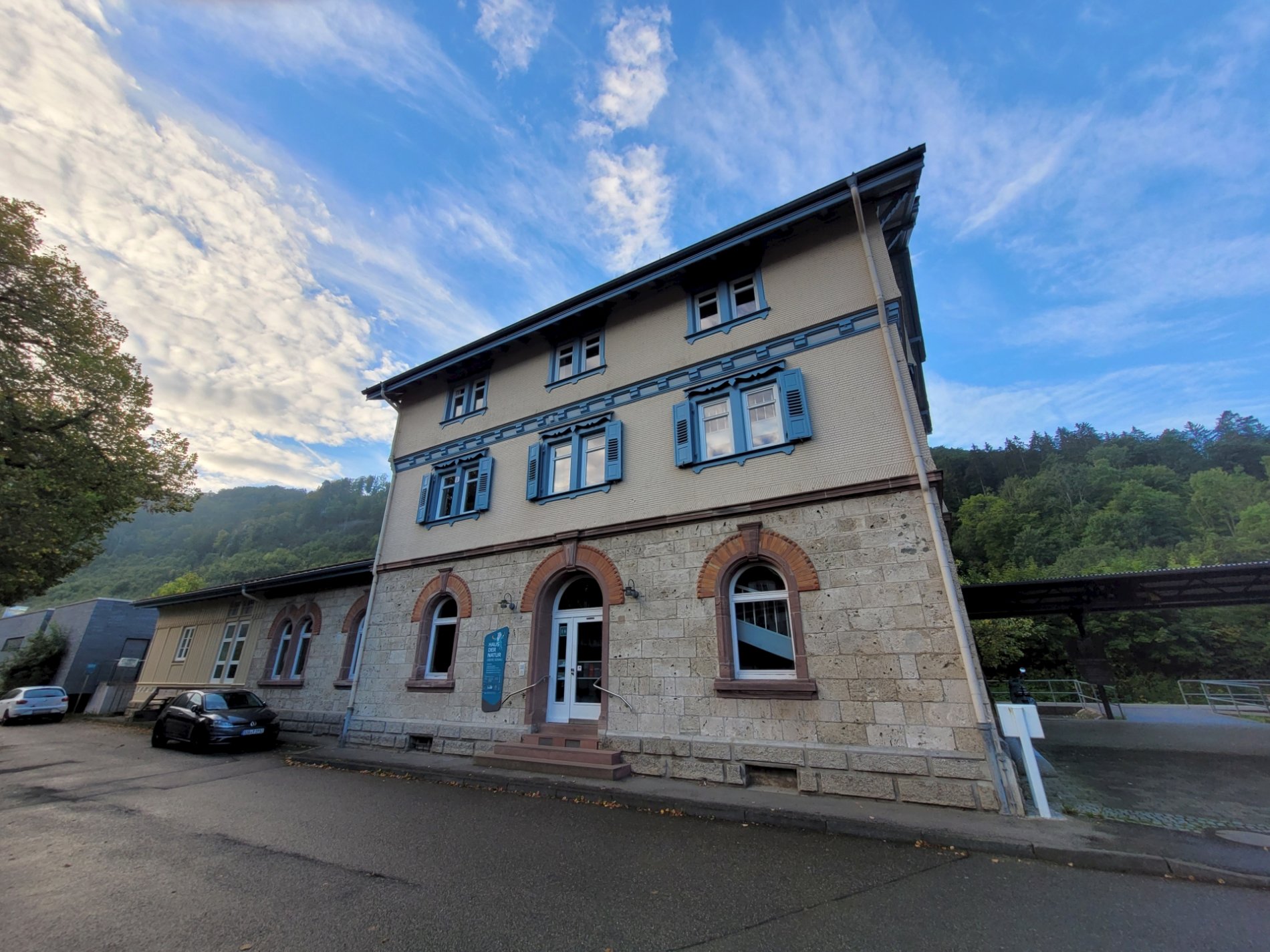 Bild vom Bahnhofsgebäude in Beuron mit drei Stockwerken, blauen Fensterläden im zweiten Stockwerk, zwei Autos davor stehend und Bäumen und blauem Himmel im Hintergrund. Das Gebäude ist aus Kalktuff.