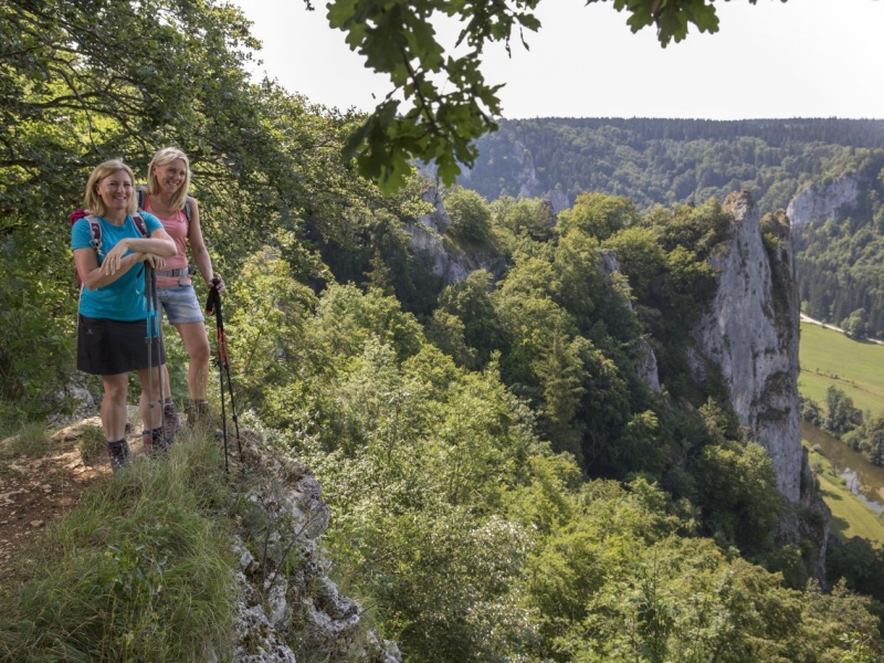 Bild von zwei Wanderinnen, die die Aussicht von oben genießen. Sie sind umgeben von Bäumen und Felsen