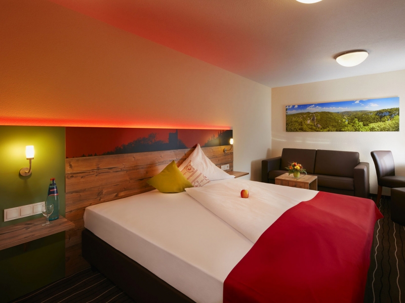 Themenbild von einem Hotelbett in einem modernen Zimmer mit brauner Ledercouch und roter Hintergrundbeleuchtung am Bett