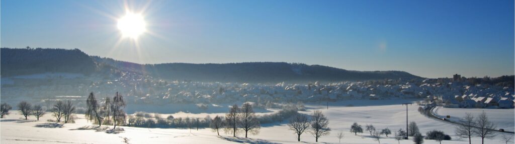 Bild von einer verschneiten Winterlandschaft, mit Wohnsiedlung im Hintergrund und strahlender Sonne