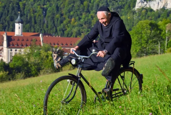 Bild von Mönch, der auf einer Wiese Fahrradfährt und die Beine von sich streckt. Im Hintergrund sieht man ein Kloster