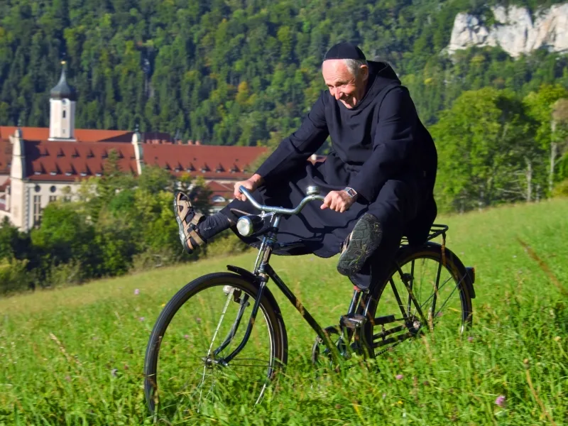 Bild von Mönch, der auf einer Wiese Fahrradfährt und die Beine von sich streckt. Im Hintergrund sieht man ein Kloster