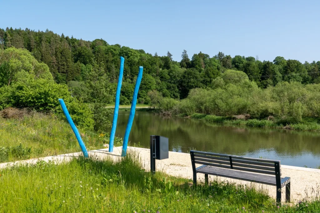 Bild vom Donauuferpark in Immendingen mit einer Parkbank am Wasser und ringsherum stehen Bäume und links im Bild sind drei hellblaue Holzstehlen als Skulptur zu sehen