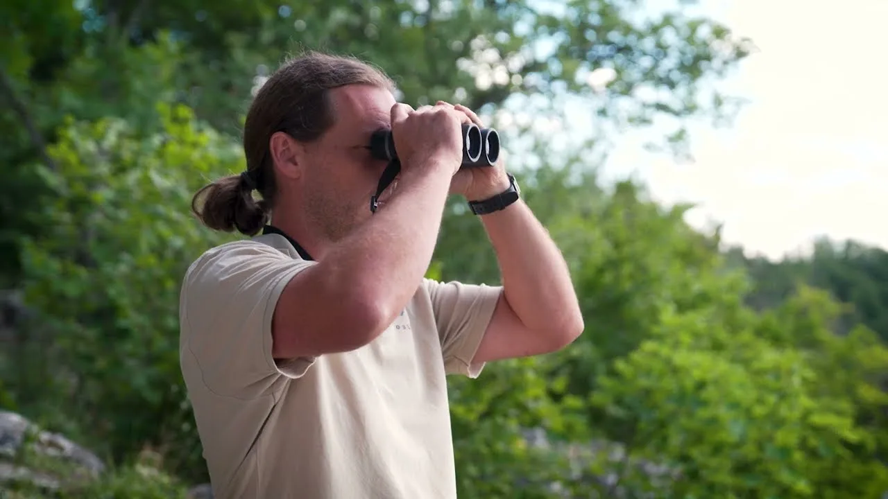 Bild von einem Mann, der vor Bäumen stehend durch ein Fernglas schaut