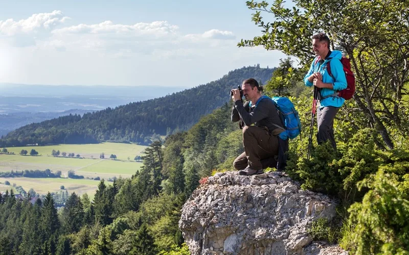 Bild von zwei Wanderern, der eine sitzt auf einem Felsen und fotografiert mit einer Kamera und der andere steht dahinter mit Wanderstöcken