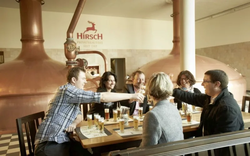 Bild von einer Bierverköstigung vor Braukesseln der Hirsch Brauerei, mit sechs Personen an einem Tisch, die sich zuprosten