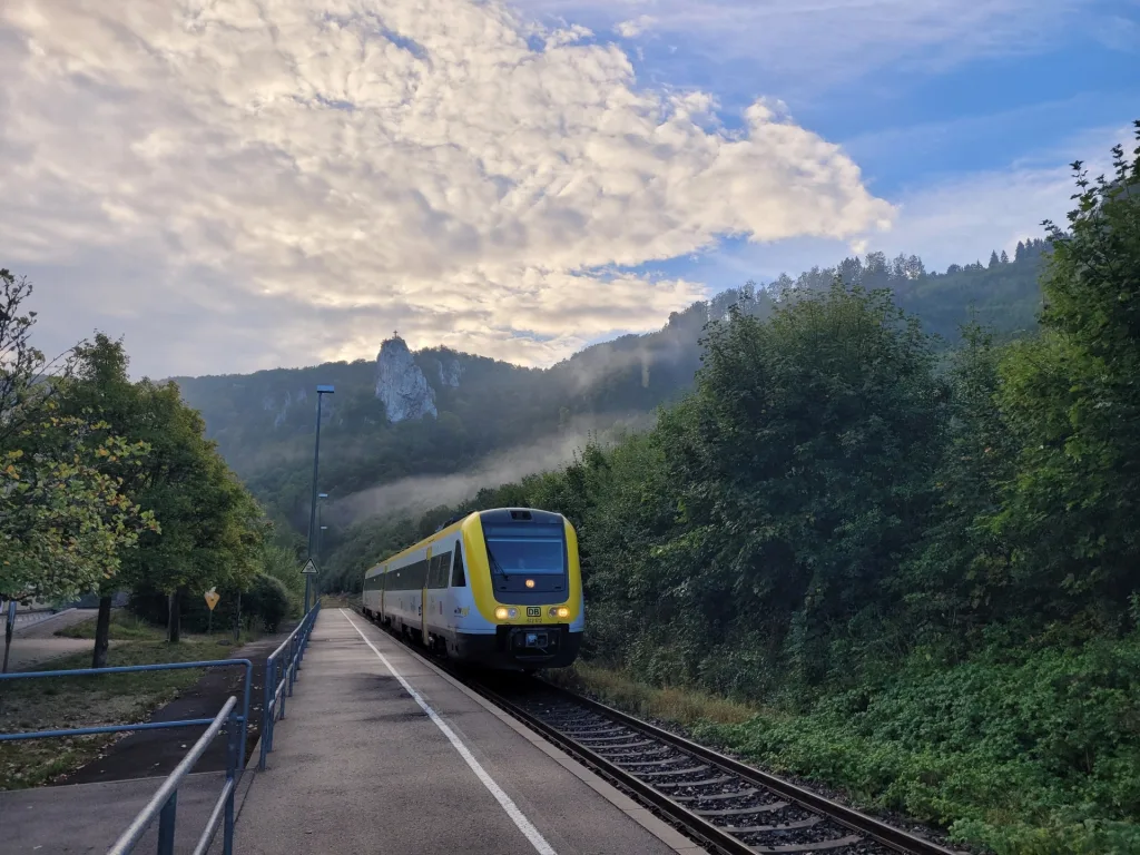 Bild eines einfahrenden Zugs in Beuron am Bahngleis. Im Hintergrund ist ein Felsen in der Höhe mit Kreuz zu erkennen