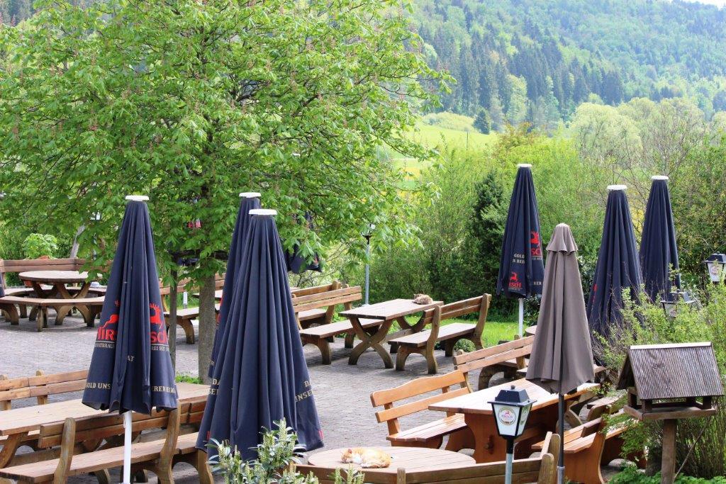 Biergarten mit Holztischen und Bänken sowie Sonnenschirmen und mit Blick ins Tal