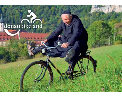 Donaubergland Postkartenmotiv mit fahrradfahrendem Mönch, der die Beine in die Luft hält und oberhalb eines Klosters in der Wiese fährt.