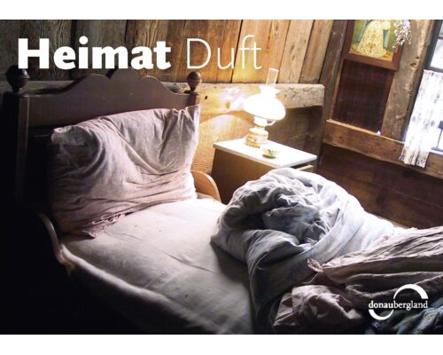 Donaubergland Postkartenmotiv mit Bett mit zurückgeschlagener Decke in einem alten mit Balken errichteten Raum.